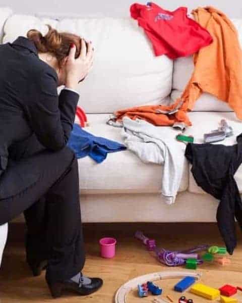 woamn sitting on a couch with her head in her hands, distraught over the mess of clothes and toys all around her