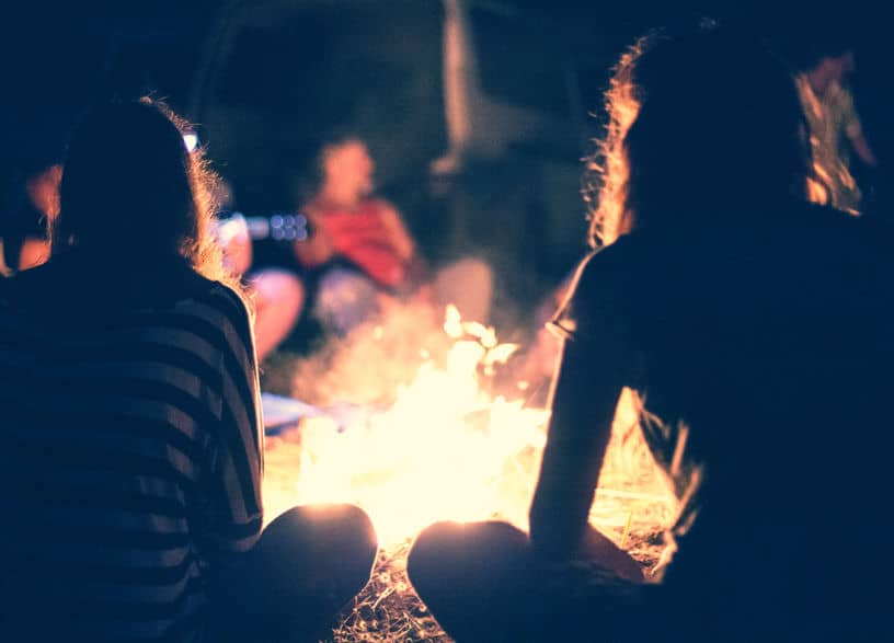 A couple of people standing around a fire. Camping Trips Can Make Fun Frugal Summer Vacations