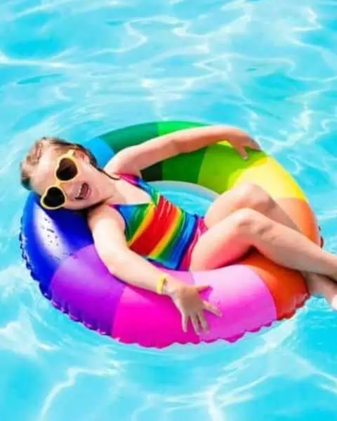 A child wearing a rainbow swimming suit floating on a rainbow inflatable in the pool.