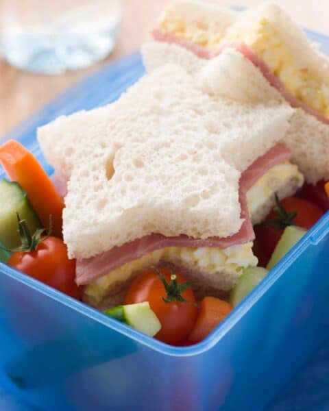 Star-shaped sandwich with ham and egg filling in a blue lunchbox, accompanied by cherry tomatoes and sliced cucumber and carrot—perfect for School Lunches Kids Love.