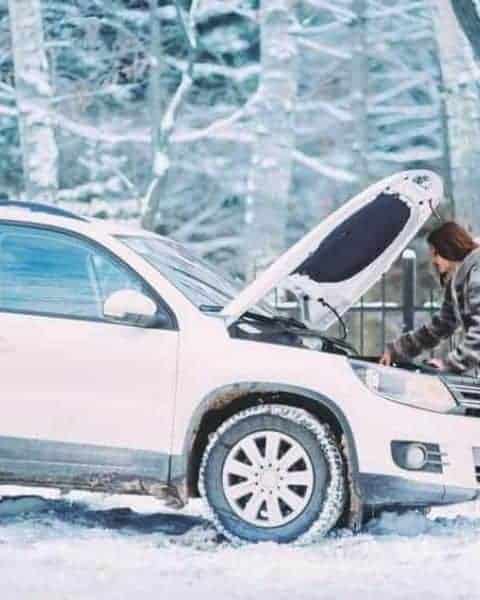 A woman with the hood of her car lifted in the middle of the snow. She is working on her car.