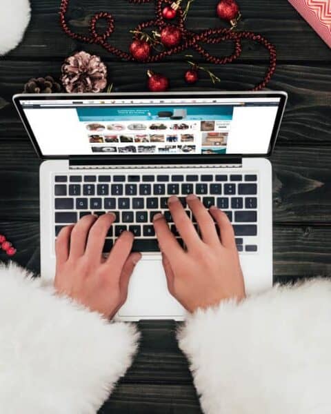 A woman wearing Christmas sweater is shopping on her laptop for holiday deals.