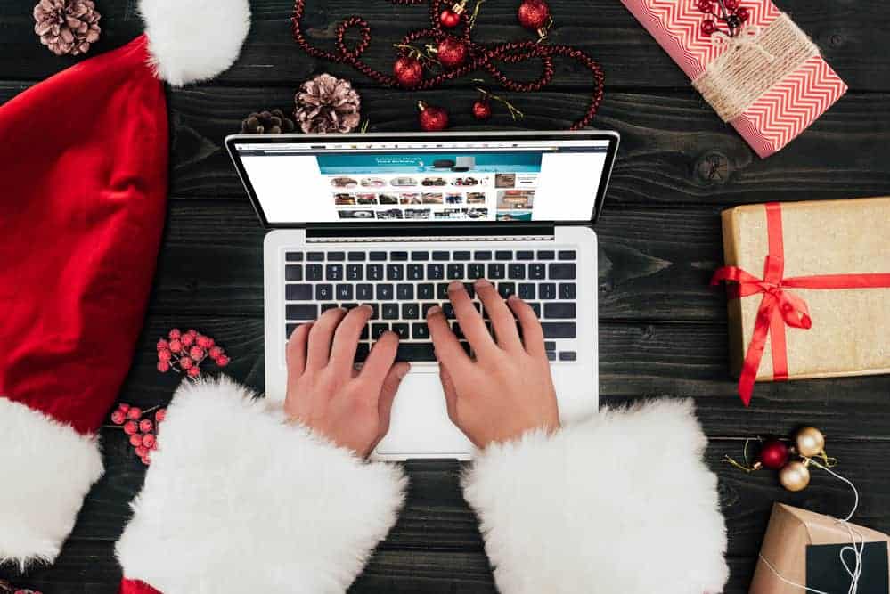 A child wearing a Santa coat while typing on a laptop and surrounded by Christmas presents and a Santa hat.
