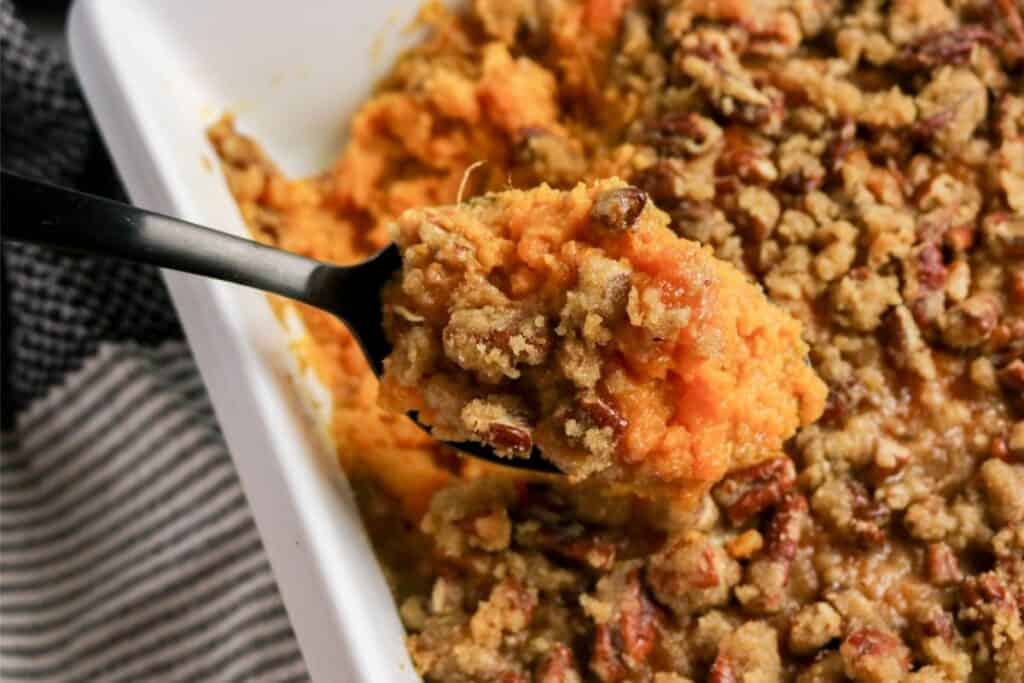 A serving spoon lifts a portion of the best sweet potato casserole recipe topped with crumbly pecan streusel from a white baking dish.