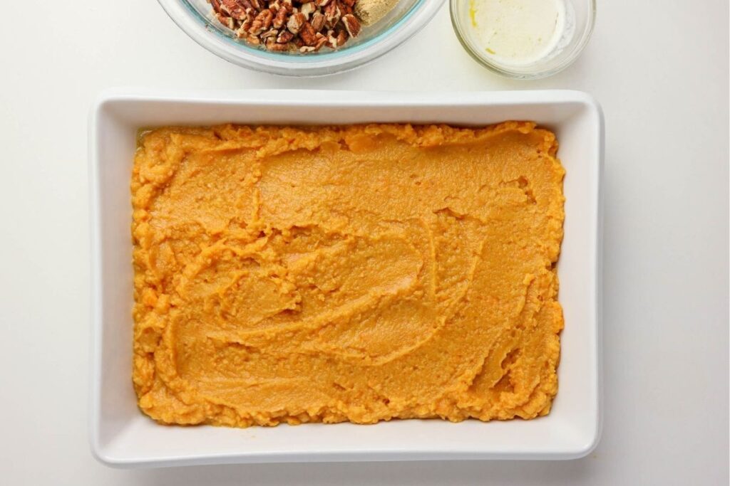 A white baking dish filled with smooth sweet potato mixture, alongside a bowl of chopped pecans and melted butter—everything you need to create The Best Sweet Potato Casserole Recipe on a bright white surface.