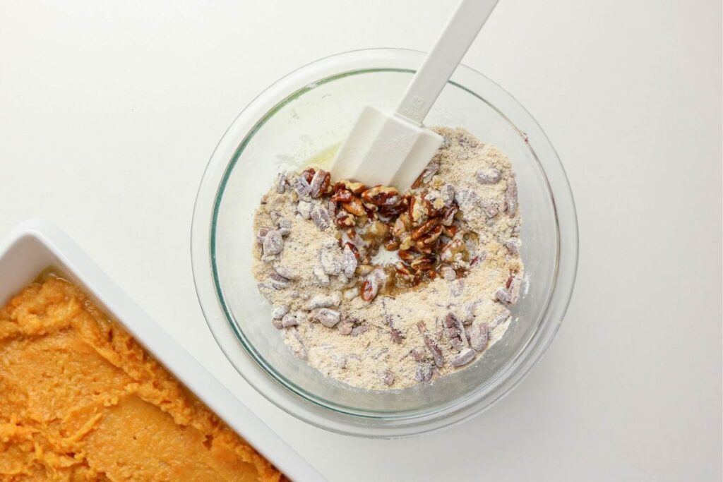A glass bowl with flour, chopped pecans, and a spatula sits beside a baking dish filled with an orange mixture on a white surface—ingredients ready for The Best Sweet Potato Casserole Recipe.