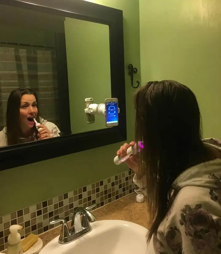 A woman brushing her teeth in front of a mirror with a rose gold Oral B 8000 toothbrush. 