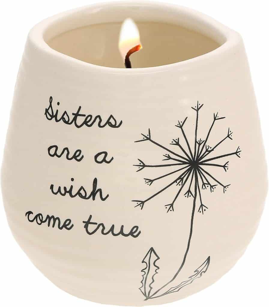 Cream-colored candle holder with a dandelion drawing, glowing candle, and the words “Sisters are a wish come true.”.