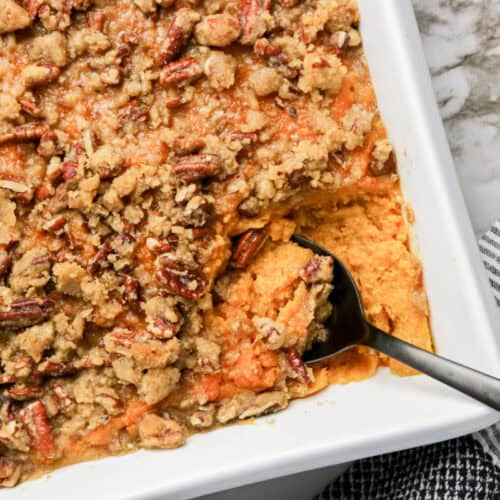 A baking dish filled with The Best Sweet Potato Casserole Recipe, topped with pecan crumble, sits ready to serve with a spoon and a generous portion already scooped out.