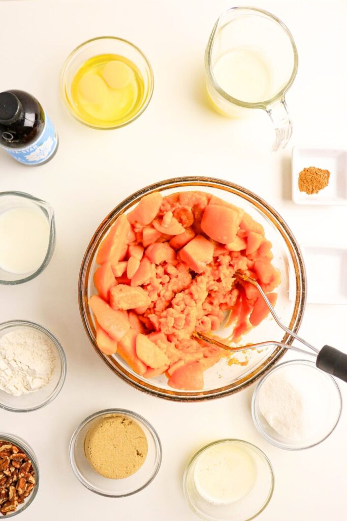 Overhead view of The Best Sweet Potato Casserole Recipe: mashed sweet potatoes in a bowl surrounded by measured ingredients like eggs, milk, sugar, flour, spices, pecans, brown sugar, and vanilla extract.