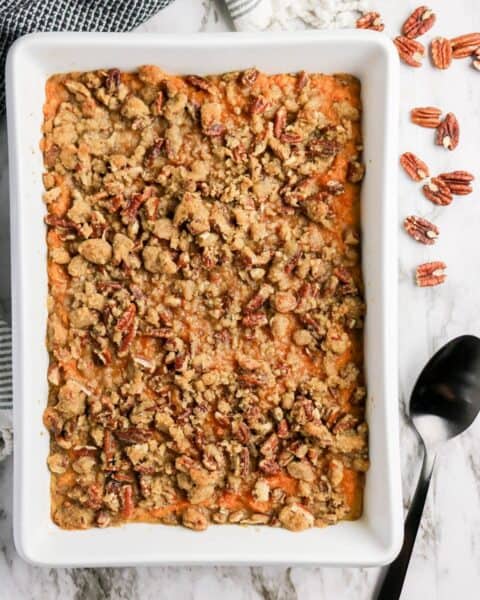 The Best Sweet Potato Casserole Recipe features a baked sweet potato casserole topped with a crumbly pecan streusel in a white baking dish, served with a spoon and pecans on the side.