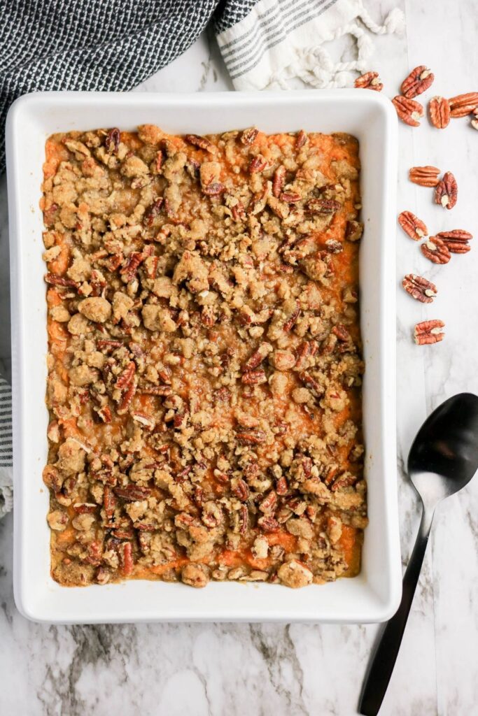 The Best Sweet Potato Casserole Recipe features a baked sweet potato casserole topped with a crumbly pecan streusel in a white baking dish, served with a spoon and pecans on the side.