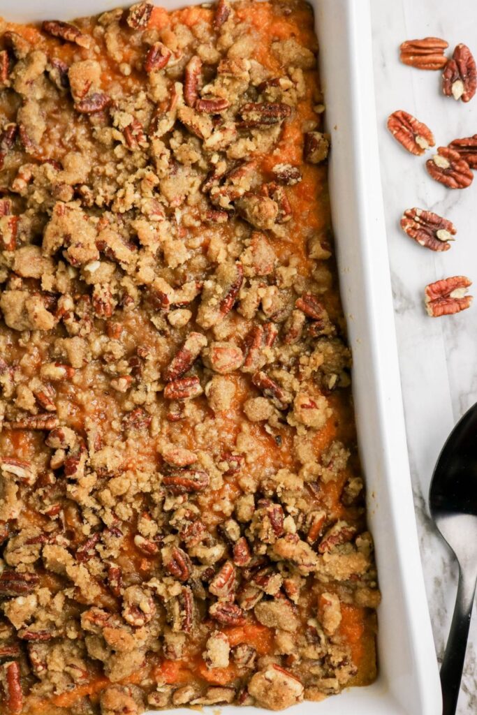 A baked sweet potato casserole topped with a crumbly pecan streusel in a white dish, with pecans scattered nearby and serving utensils on a marble surface—The Best Sweet Potato Casserole Recipe for your holiday table.