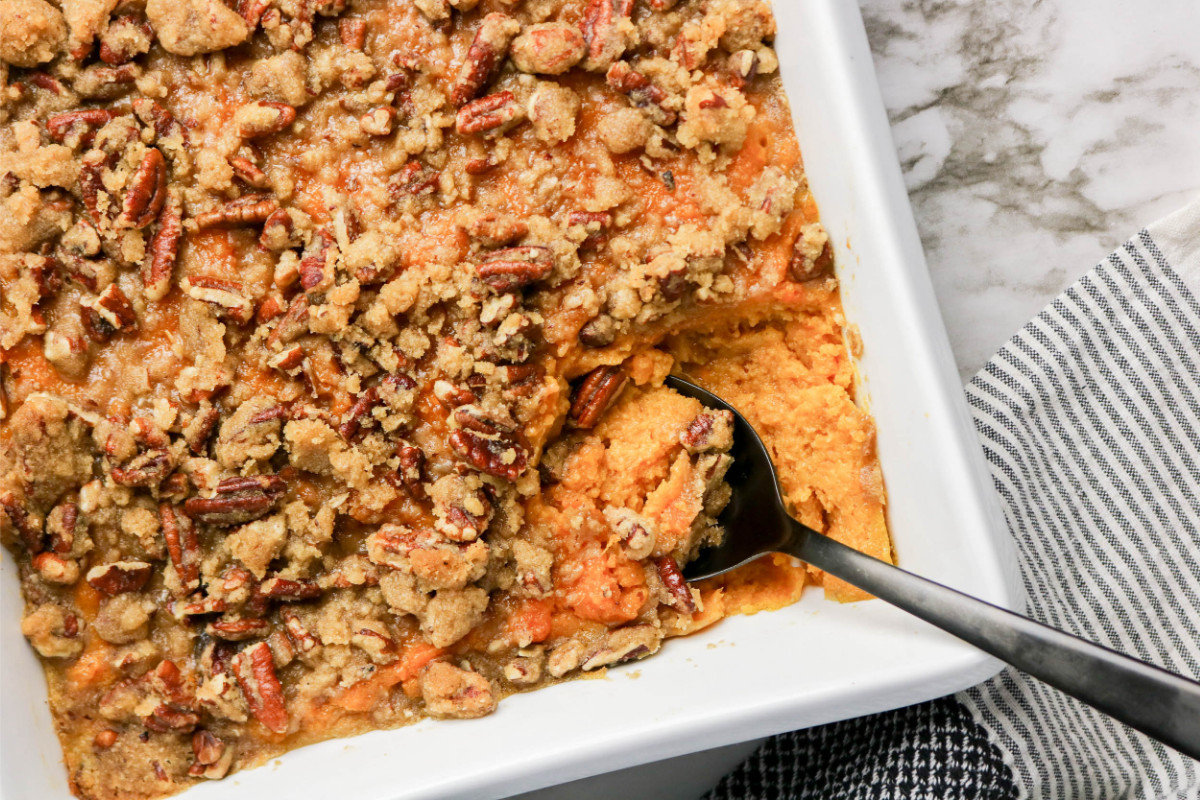A baking dish filled with The Best Sweet Potato Casserole Recipe, topped with pecan crumble, sits ready to serve with a spoon and a generous portion already scooped out.