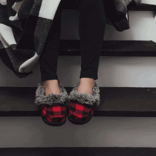 A woman sitting on the stairs wearing a pair of MUK LUKS moccasin slippers, on sale!