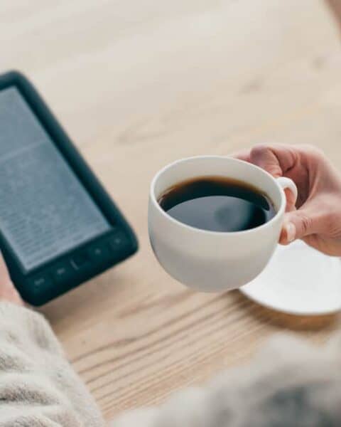 A woman drinking a cup of coffee while reading from her Kindle unlimited.