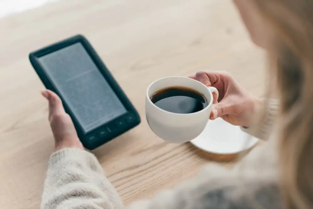 A woman drinking a cup of coffee while reading from her Kindle unlimited.