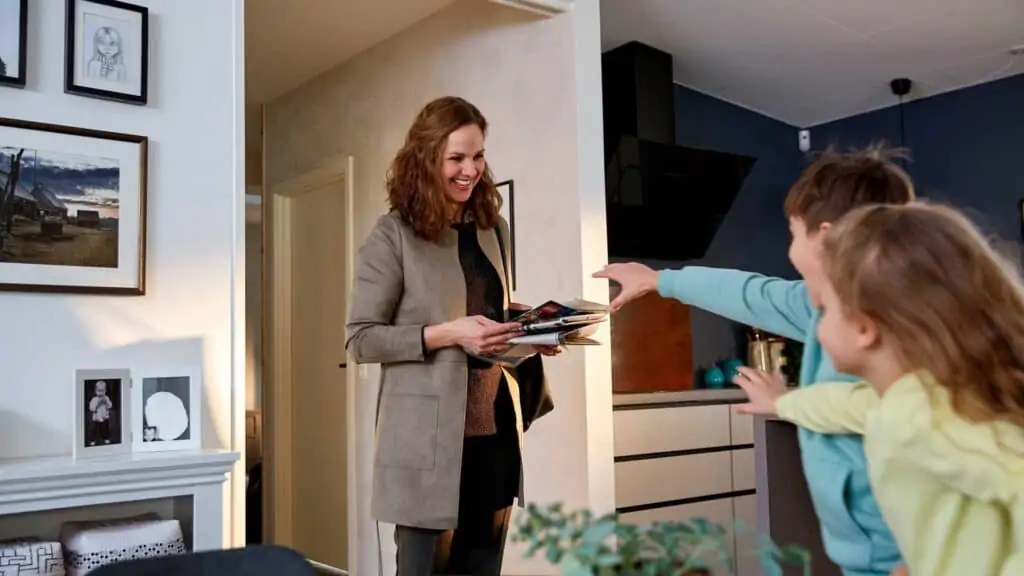 Mom walking into living room with a pile of magazines. Her children's hands are outstretched to grab their copies.