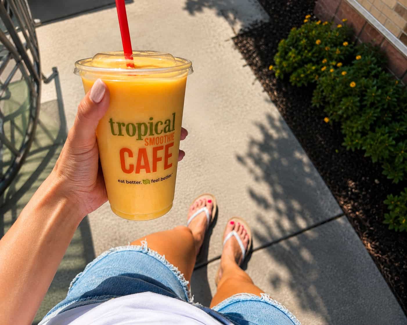 Person holding a yellow smoothie from a smoothie cafe, standing on a sidewalk in flip-flops and denim shorts, with greenery and flowers nearby.