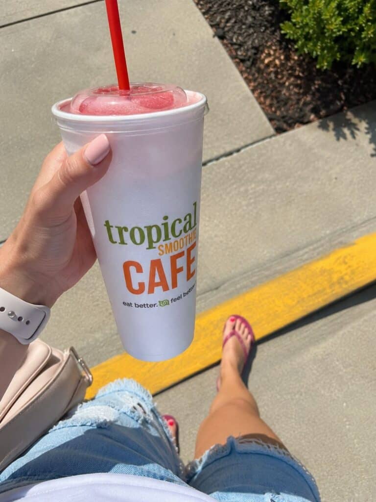 Person holding a smoothie cafe cup with a red straw, standing on a sidewalk in sandals and denim shorts.