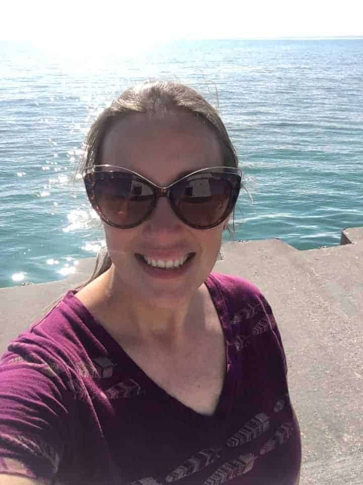 A smiling woman who is wearing sunglasses while standing in front of a body of water.