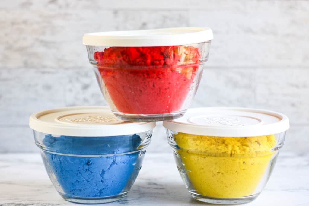 Three separate containers of red, blue, and yellow Homemade Kinetic Sand Recipe.