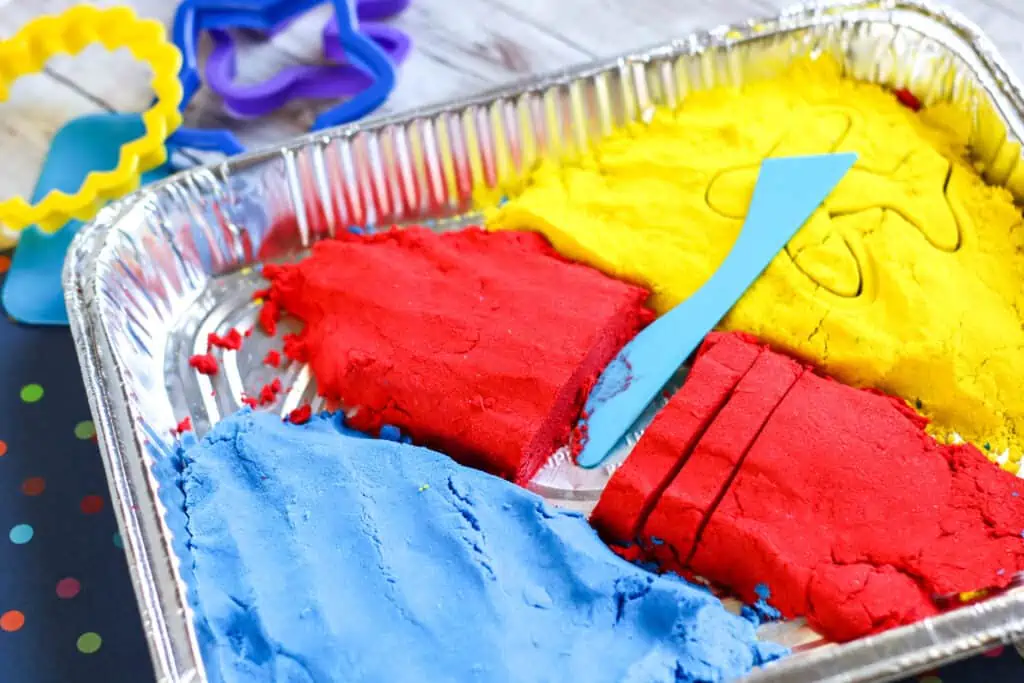 Blue, red, and yellow Homemade Kinetic Sand Recipe with cookie cutters and other play things for the sand.