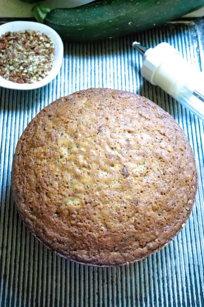 a double layer zucchini cake on a table with a zucchini and a bowl of chopped pecans.