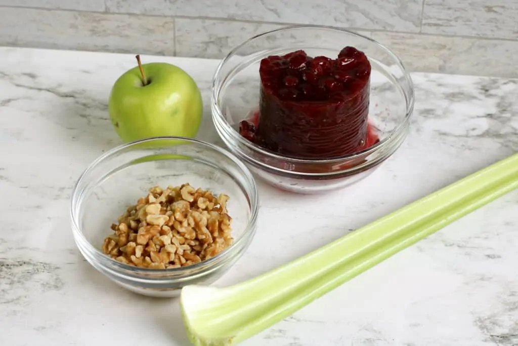 All of the ingredients to make cranberry apple salad sitting on a counter top - Granny Smith apple, walnuts, celery stalk and cranberry. 