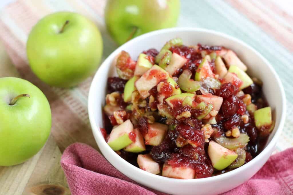 A bowl of cranberry apple walnut salad sitting on a table near three granny smith apples.