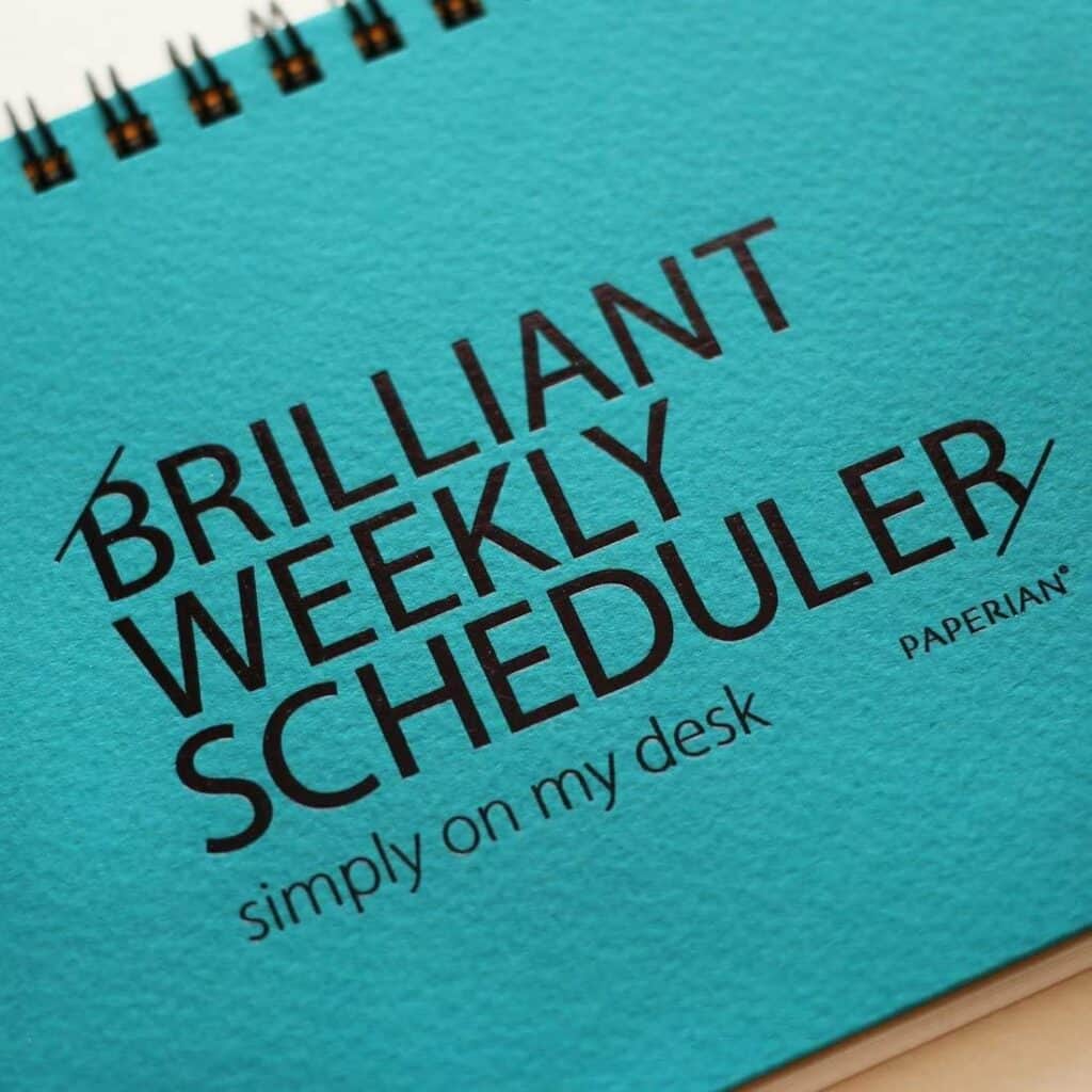 A turquoise spiral-bound planner with "BRILLIANT WEEKLY SCHEDULER simply on my desk PAPERIAN" in bold black letters—an excellent addition to your workspace and perfect for Gift Ideas for Teachers.