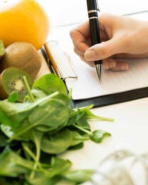A woman writing in her notebook next to kiwi's, oranges, and spinach.