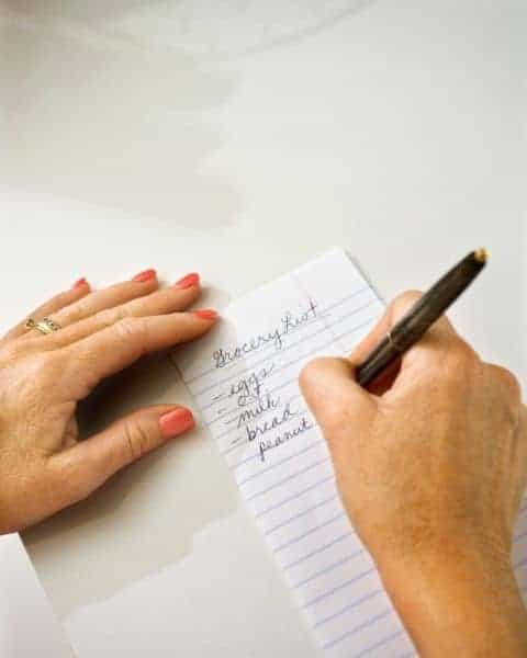 A woman making a grocery list, including eggs, milk, bread, and more food items on the list.