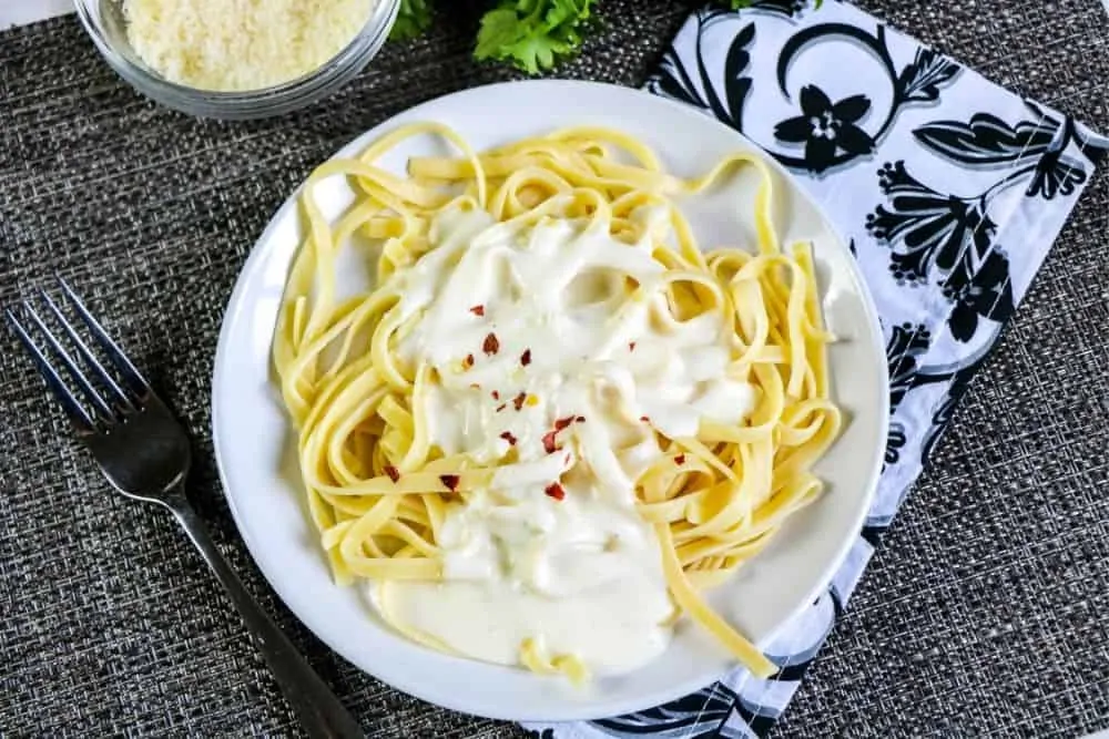 a dinner plate filled with pasta and garlic alfredo sauce sitting on a table near a bowl of grated Parmesean cheese. 