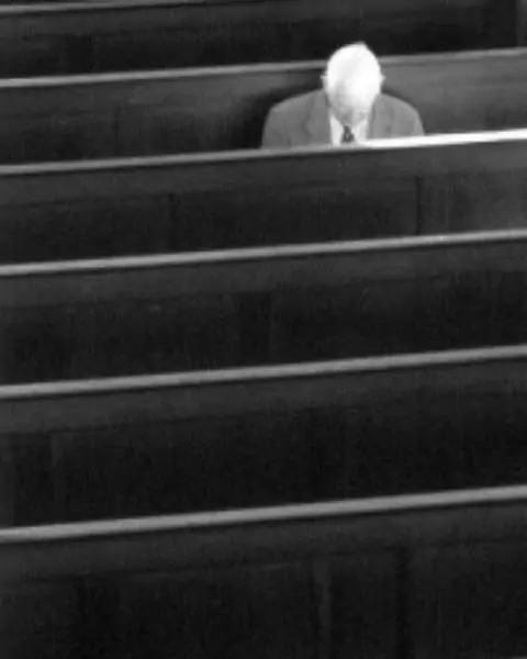 An old man sitting in a church pew by himself.