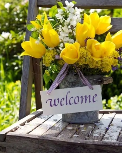 An outdoor wooden chair with a potted plant of flowers on the seat of the chair with a "welcome" sign.