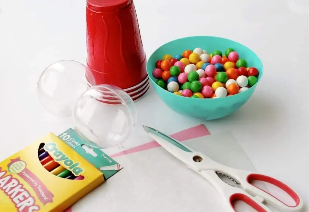 All of the items needed to make mini bubble gum machines: an empty clear ornament, red plastic cups, a bowl full of colored mini gum balls, scissors, construction paper and markers. 