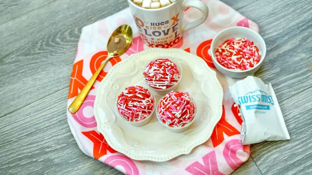 Three hot cocoa balls covered in red, white and pink sprinkles sitting on a white ceramic plate, surrounded by Swiss Miss hot cocoa packet, more sprinkles in a white bowl, a mug of hot cocoa and mini marshmallows and a spoon. 