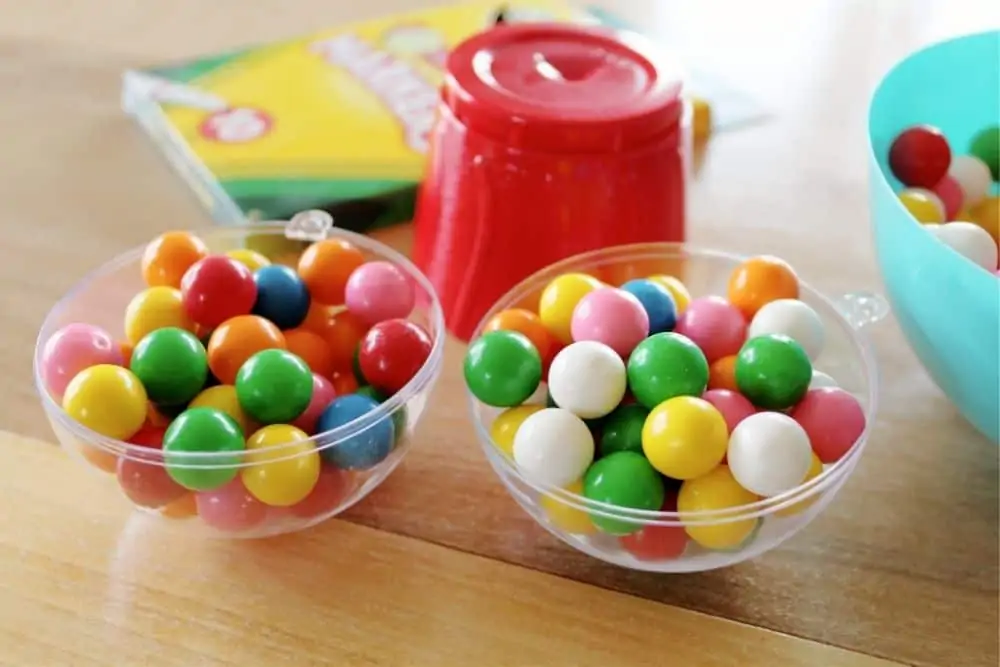 An opened plastic clear ornament sitting on a table filled with brightly colored gum balls. 