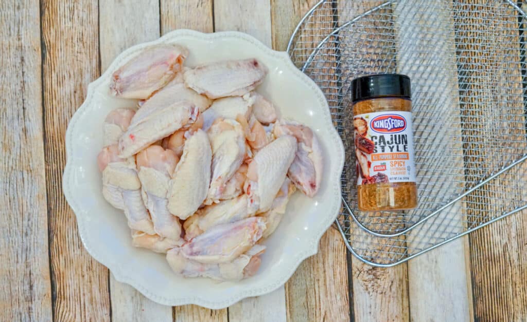 A plate filled with raw chicken wings sitting on a table next to a canister of Cajun Seasoning blend. 