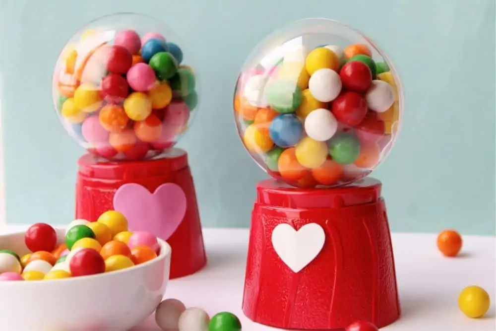 A white ceramic bowl filled with red, green, pink, orange, white and yellow mini gumballs sitting on a table next to two crafted mini gum ball machines.
