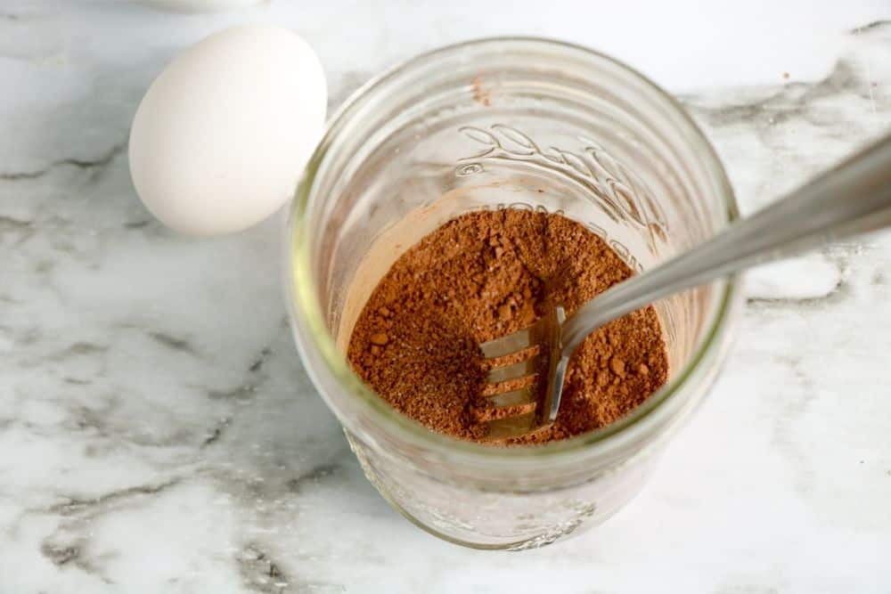 chocolate mug cake dry ingredients in a mason jar with a fork, sitting on a counter next to an egg. 