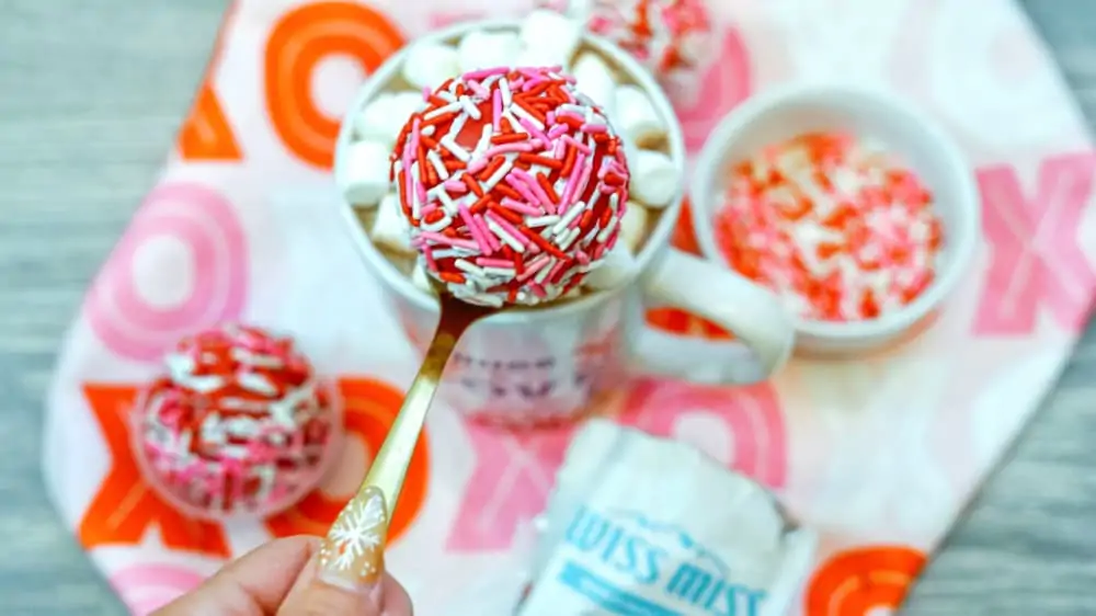 Close up photo of a completed mini hot chocolate ball covered in red, white and pink sprinkles with a mug of hot cocoa in the background.