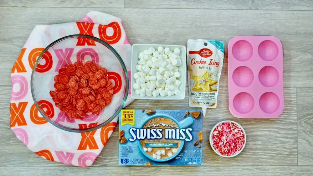 Ingredients to make Valentine's Day hot chocolate balls, including red candy melts, mini marshmallows, white icing, silicone mold, Swiss Miss hot chocolate and Valentine's Day sprinkles. 