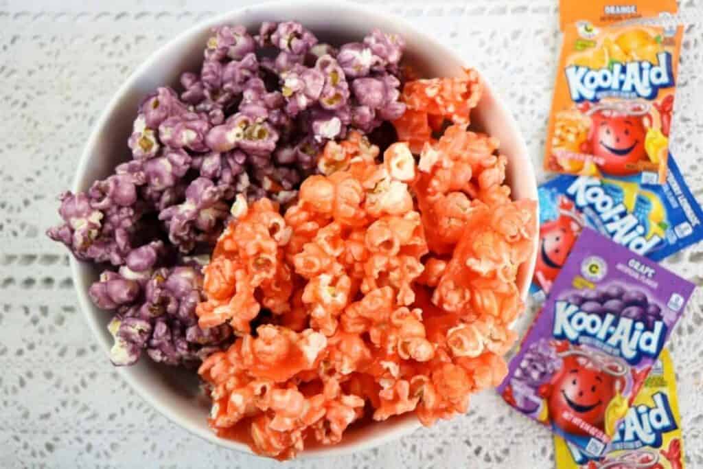 A cowl filled with purple and orange colored popcorn sitting next to several packets of Kool Aid drink mix. 