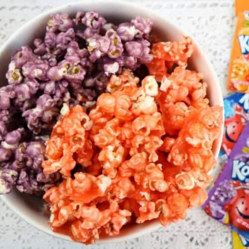 A cowl filled with purple and orange colored popcorn sitting next to several packets of Kool Aid drink mix.