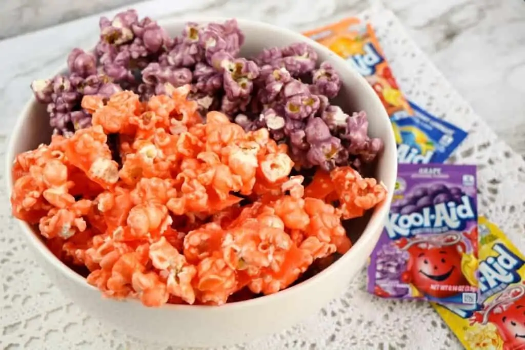 Orange and purple colored popcorn in a white bowl, sitting on a table next to packets of drink mix. 