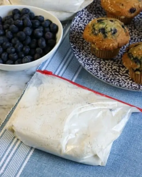 muffin mix in a ziplock bag sitting on a table with a bowl of blueberries and a plateful of blueberry muffins.
