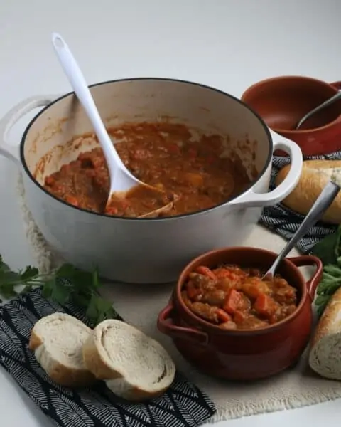 A pot of beef stew sitting near a bowl of stew with homemade bread nearby.