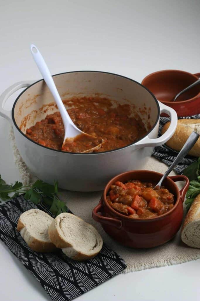 A pot of beef stew sitting near a bowl of stew with homemade bread nearby.